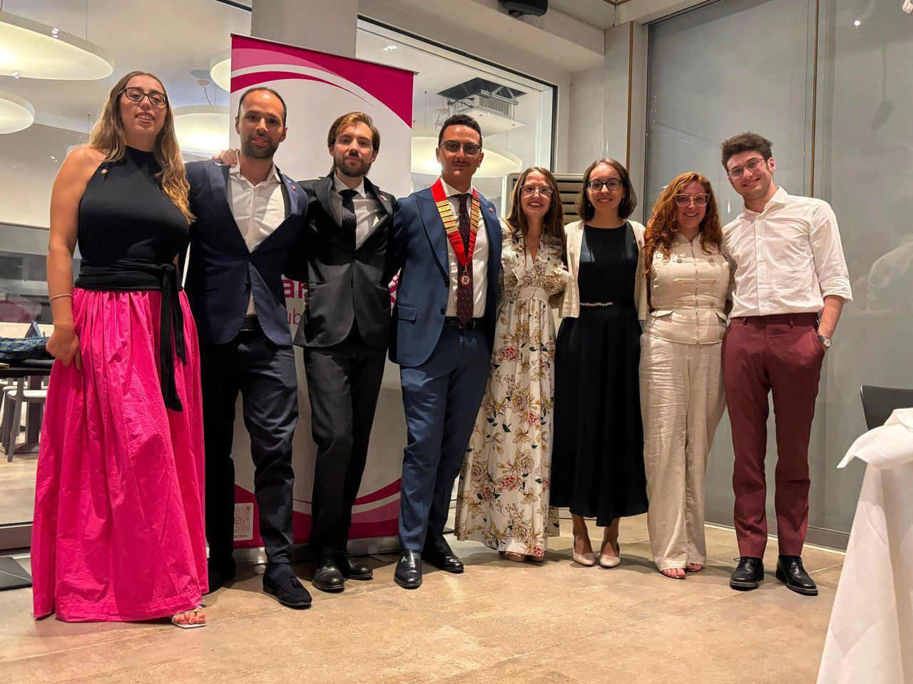 Foto di gruppo al Passaggio di Consegne: otto persone in abito elegante in posa davanti a un banner.